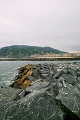 Fototapeta premium Vertical view of massive grey stones of a breakwater in the foreground, with the sea water, city skyline, and a prominent green hill under a cloudy sky in San Sebastian, Basque Country, Spain territor