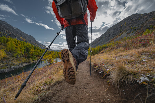 A low angle shot of a hiker with a backpack and trekking poles actively walking uphill on a mountain trail beside a river, surrounded by vibrant autumn trees and dramatic clouds.