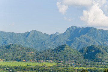 Obraz premium A vibrant rural settlement sits at the base of lush, forested mountains under a bright blue sky dotted with scattered clouds in northern Vietnam. The landscape features green rice fields, rolling