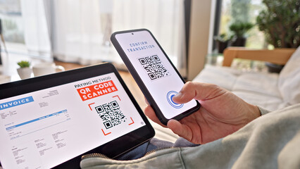 Fototapeta premium A person uses a smartphone to scan a QR code on a tablet for a contactless payment in a cozy indoor setting, highlighting modern digital transactions and convenience in online shopping and finance.