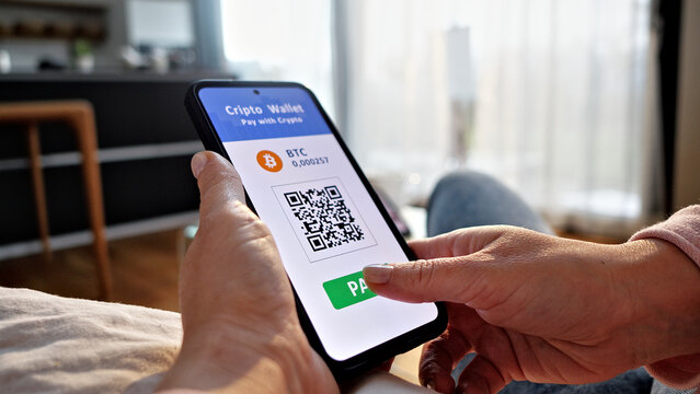A person holds a smartphone with a cryptocurrency wallet app showing a QR code for contactless payment. The cozy indoor setting suggests online shopping or digital finance transactions.