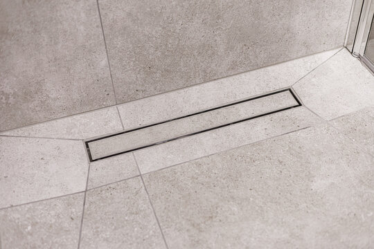 Close-up of a sleek linear drain integrated into a light gray stone-tiled shower floor. Clean, geometric lines emphasize modern bathroom design
