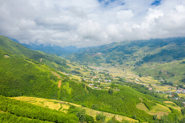 Fototapeta premium Wide panoramic scene of lush green hills and layered rice terraces surrounding a rural valley near Sapa, Vietnam. Cloud-filled skies cast soft light over the vibrant landscape and distant village.