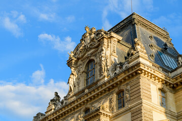 Obraz premium Warm evening sunlight illuminates the elaborate rooftop statuary and decorative stonework of the Louvre palace in Paris, France. The intricate renaissance details and sculpted figures stand out