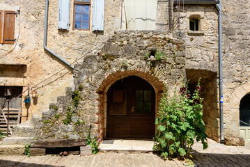 Fototapeta premium Traditional stone building in La Couvertoirade featuring an arched wooden doorway, weathered exterior stairs, and tall hollyhock flowers growing beside a sunlit cobblestone street.