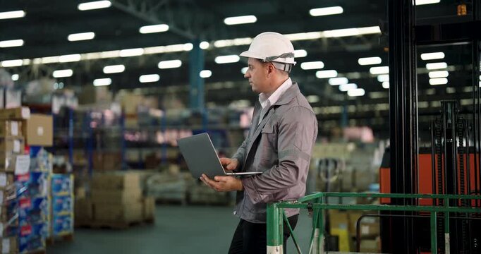 caucasian male warehouse staff working with laptop near stacked boxes checking stock status beside tall pallet racks in industrial factory storage area under bright ceiling lights