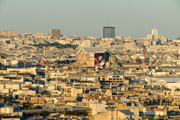 Wide view over Paris rooftops with a mix of classic and modern buildings, highlighted by soft golden sunlight and a clear sky. Urban density and architectural variety create a textured cityscape.