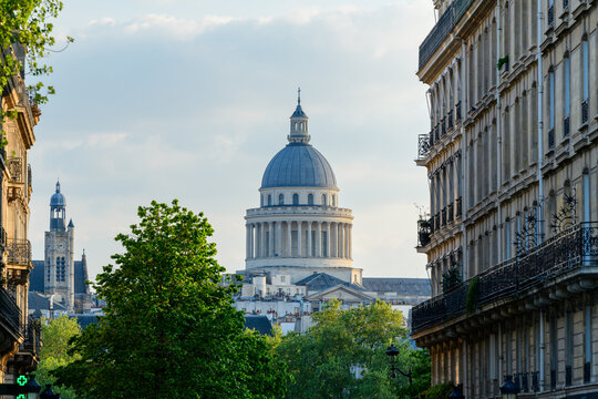 The grand dome of the Pantheon rises above leafy trees and is framed by ornate Parisian apartment buildings under a soft, partly cloudy sky.