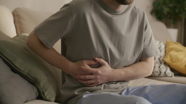 Man sitting on sofa and holding abdomen with both hands, bending forward while suffering from stomach pain at home