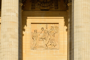 Sunlit neoclassical stone relief panel framed by tall fluted columns on the Pantheon facade in Paris, featuring detailed sculptural figures and warm golden tones.