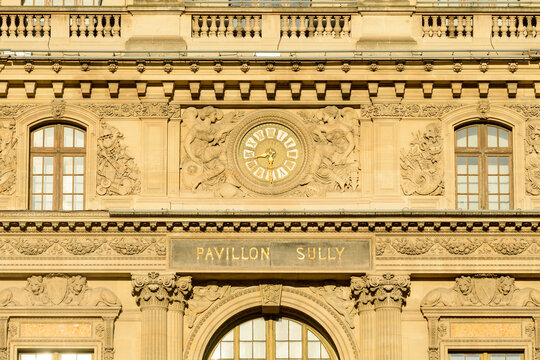 Frontal view of the Pavillon Sully at the Louvre in Paris, France, featuring a central gold-accented clock surrounded by detailed classical stone bas-reliefs and ornate architectural motifs. Warm