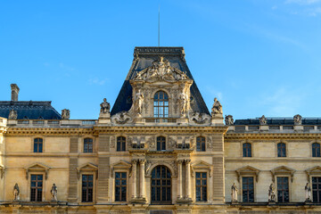 Obraz premium Symmetrical frontal view of the Louvre palace's central pavilion in Paris, France, featuring detailed classical statues and elaborate stone reliefs beneath a slate mansard roof. The historic facade is