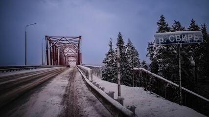 Fototapeta premium Snowy bridge crossing in northern Russia. Icy road leads through a red steel bridge surrounded by winter forest, with a road sign covered in snow and a cold, moody sky.