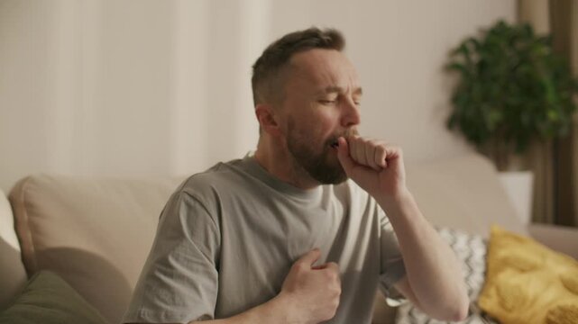 Man sitting on couch at home, coughing with his hand on chest as experiencing sudden choking episode or symptoms of respiratory illness