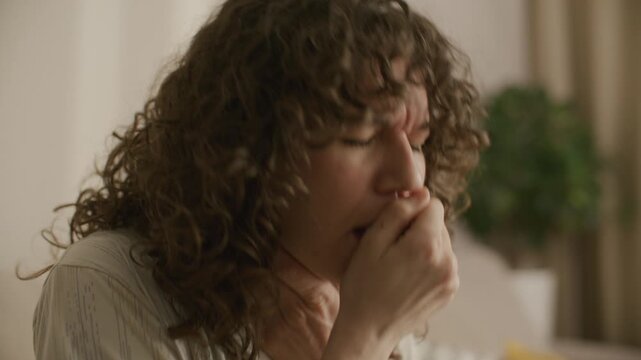 Sick woman with curly hair sitting in living room, coughing into fist and touching her neck while suffering from respiratory illness