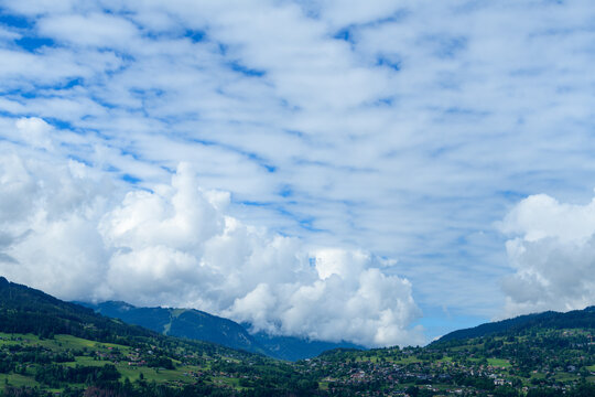 Sweeping view of dramatic cumulus clouds drifting above the lush green hills and scattered village rooftops of Combloux, framed by distant alpine mountains under a vibrant blue sky.