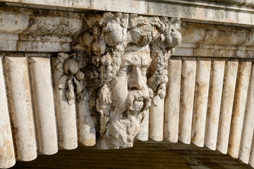 Close-up of a detailed stone face and fruit garland carving on Le Pont Notre Dame bridge. Warm sunlight enhances the aged texture and intricate craftsmanship of the sculpted masonry.