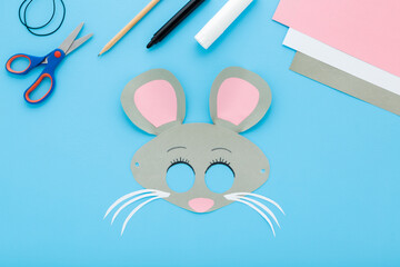 Created gray mouse face mask. Scissors, marker, application paper, elastic string on light blue table background. Pastel color. Closeup. Little child handcraft. Top down view.