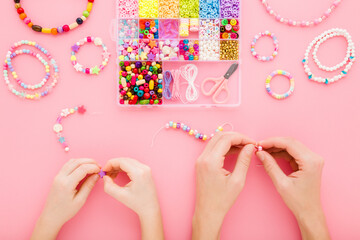 Mother and little child hands creating beaded jewellery from different colorful plastic parts on light pink table background. Pastel color. Closeup. Point of view shot. Flat lay. Top down view.
