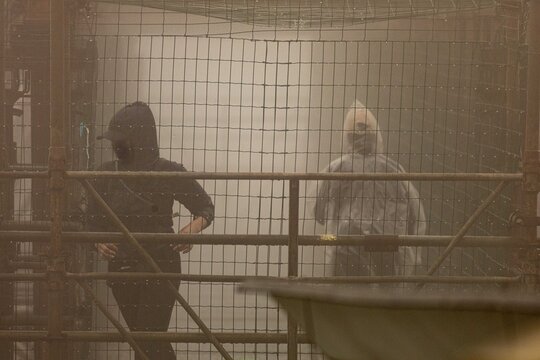 Pessoas usando capa de chuva atravessando passarela cercada de grade. 