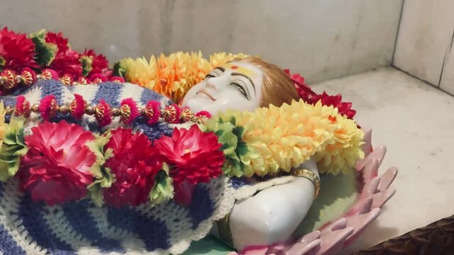 Child Krishna resting peacefully and decorated with colorful flower garlands and a knitted blanket inside a temple.