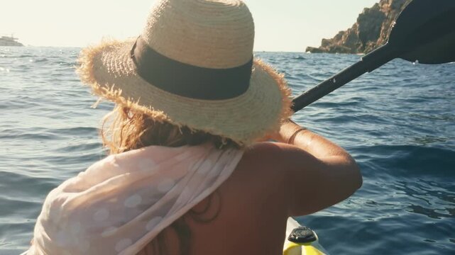 A lone kayaker in a straw sunhat and polka dot scarf paddles toward a rocky headland on the Adriatic in Montenegro, warm late afternoon light sparkles on water.