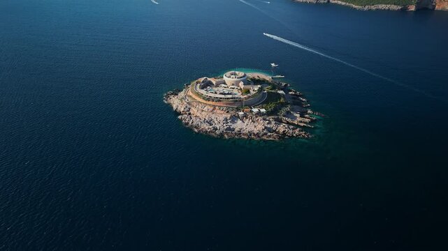 Aerial daylight view of Fortress on Lastavica, Montenegro. Boats move, wakes streak the deep Adriatic, turquoise shallows meet the rocky shore.