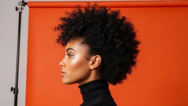 Profile view of a young woman with voluminous afro hair and glowing skin, wearing a black turtleneck against a vibrant orange studio background.
