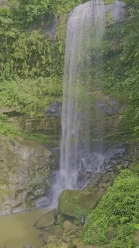 Vertical panning shot of a majestic waterfall cascading down rocks. Camera pans right to left, following the water stream flow from the top drop to the river below in a lush green forest.