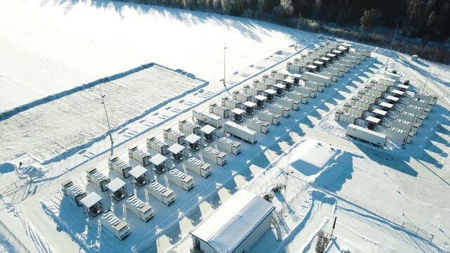 Aerial view of a large battery energy storage facility in winter, showing containerized power units casting long shadows on snow with forest surroundings and smooth orbital motion.