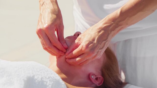 Tight close up shows rhythmic pinching and circular motions on a woman's jaw and cheeks in Montenegro, with white linens, warm light, and shallow depth of field.
