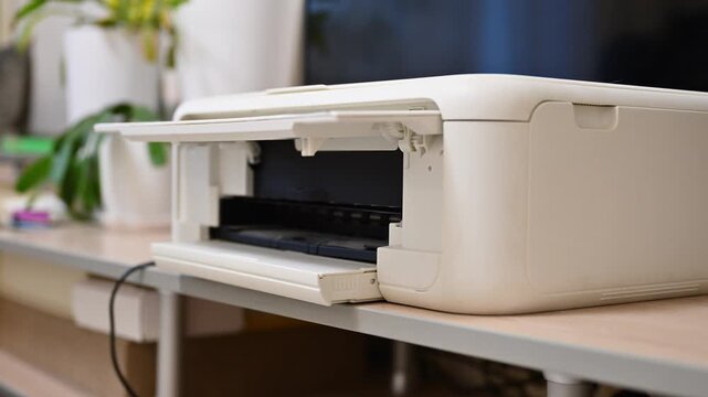 Inkjet printer actively printing documents on a desk in a modern home office. Open paper tray, everyday office workflow, home printing process, technology and remote work concept.