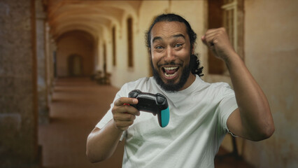 Man holds a game controller and pumps fist in a sunlit old building corridor after a big multiplayer win, mouth open in cheer  gaming excitement. © Krakenimages.com