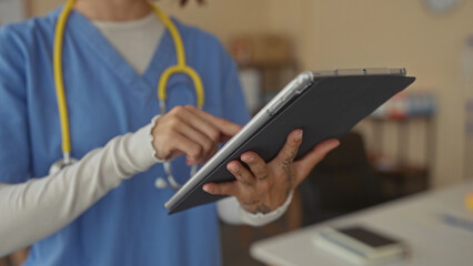 Obraz premium Woman nurse in blue scrubs taps digital tablet with tattooed hand near yellow stethoscope in clinic building; professionalism focus.
