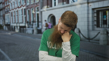 Fototapeta premium Man in green volunteer shirt and white long sleeve undershirt resting head on fist on urban street; contemplation.