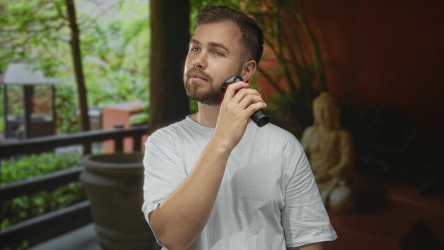Man shaves beard with electric razor while holding the razor to his jaw in a hotel building; grooming confidence.