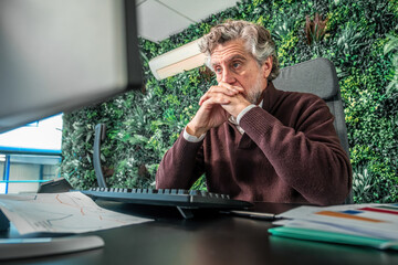 Senior businessman working on computer at an office desk, deeply focused and thinking in a modern workspace with a green wall