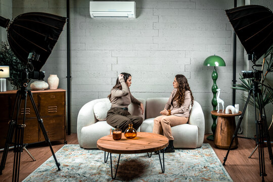Two women sitting on a sofa, conversing during a podcast or interview recording in a professional studio setup