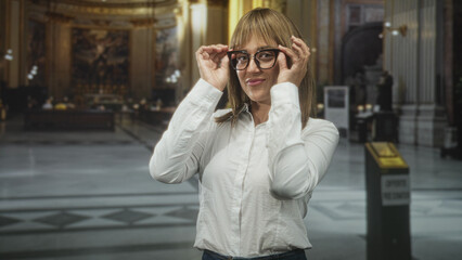 Fototapeta premium Woman adjusts glasses with hands and points with index finger inside ornate building; playful curiosity.
