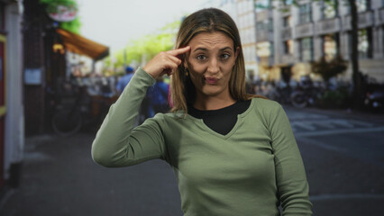Fototapeta premium Woman with finger to temple and raised hand in green top standing on a busy city street near cafe and parked bicycles; skepticism.