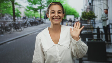 Fototapeta premium Woman smiling and waving hand on city street beside parked bicycles and trees, wearing white blouse and gold earrings; friendly greeting.