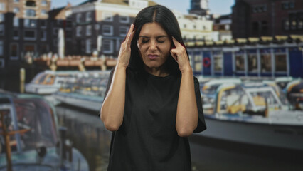 Fototapeta premium Woman pressing temples with hands, eyes closed and grimacing, visible forehead and fingers, wearing a black t shirt by canal boats on a street with buildings; stress fatigue tension.