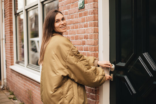 Happy brunette woman in gold bomber jacket locking or open red brick house door, leaving or back home, smiling, confident, lifestyle, city, everyday routine, urban life, morning activity