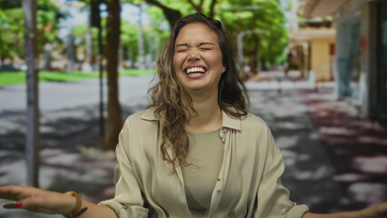 Fototapeta premium Hispanic woman smiling outdoors on a sunny street, wearing casual clothing, embodying happiness and joy amidst urban trees and vibrant surroundings.