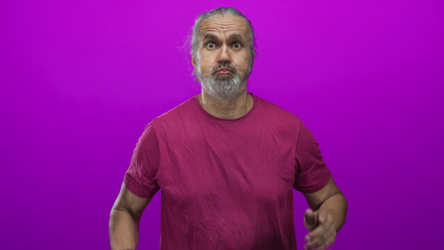 Man with hoary long hair and gray beard points fingers to cheeks in studio with magenta backdrop; playful expression.