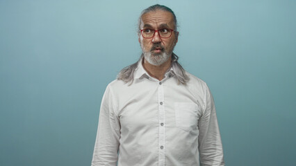 Obraz premium Man with long gray hair and beard wearing red glasses and white shirt purses lips in light blue studio; surprise and quiet introspection.