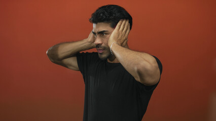 Young hispanic man covers ears with hands against orange studio wall; sound sensitivity overload anxiety.