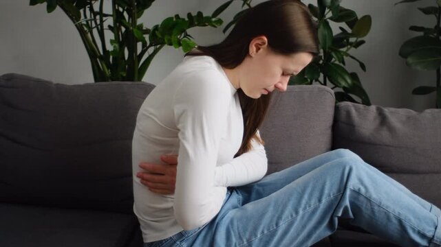 Sick young woman in pain holding belly stomach feeling hurt abdomen ache gastritis pancreatitis symptoms. Sad female sit on couch suffering from indigestion flatulence abdominal problem