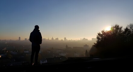 Fototapeta premium A man stands on a hill overlooking a city skyline during a serene sunset.