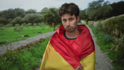 Man wrapped in red and yellow flag on a rural street holding flag with hands and looking dejected; defeat disappointment.